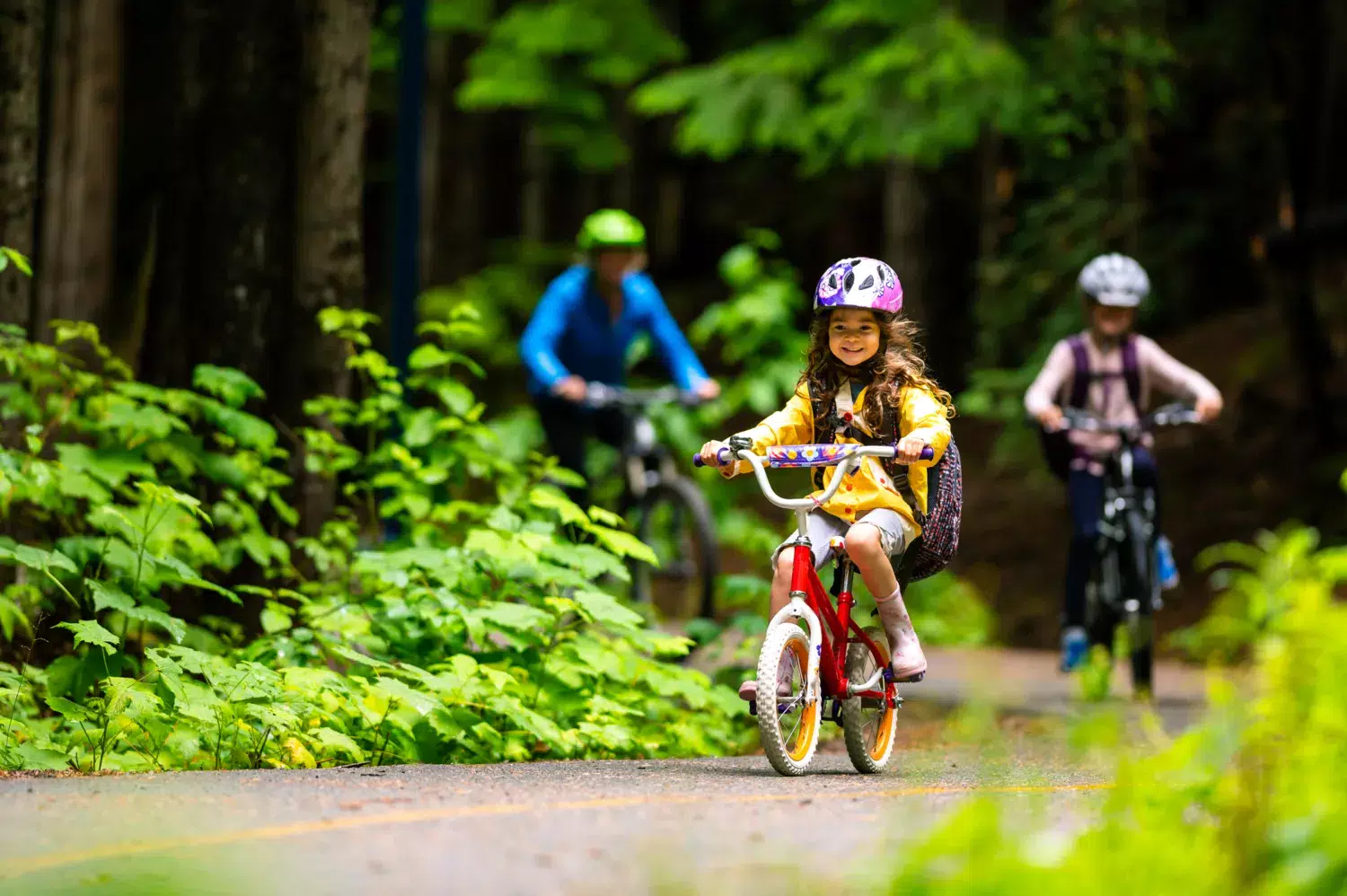 En liten flicka cyklar i skogen med sin mamma och ett syskon. Hon har cykelhjälp på sig.