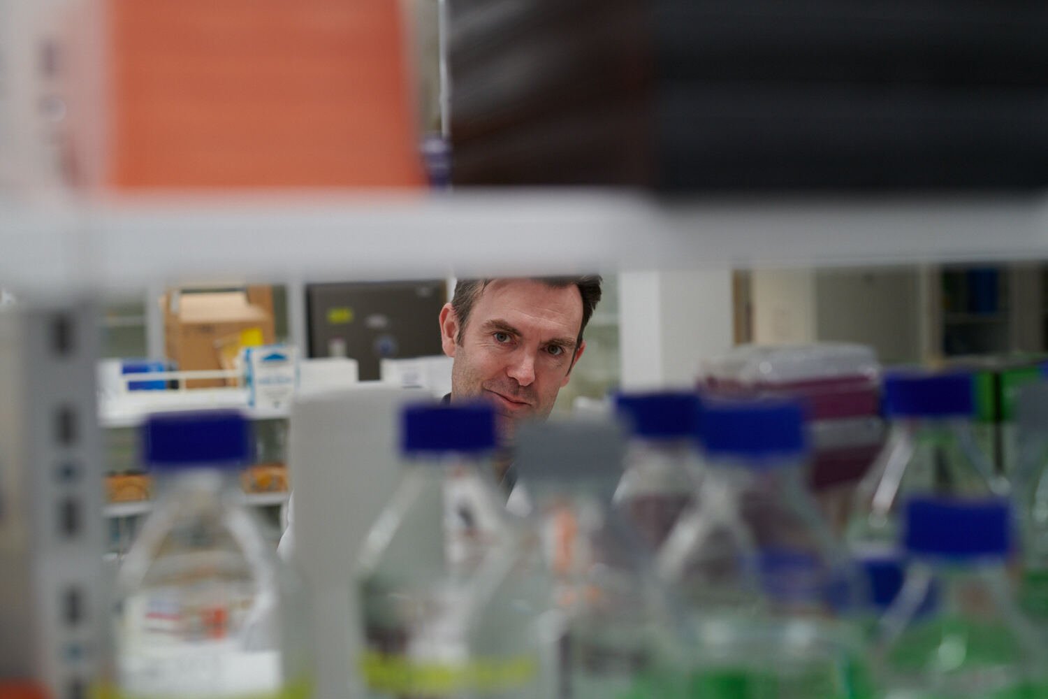 Portrait of Gerald McInerney looking between bottles and apparatus in the lab.