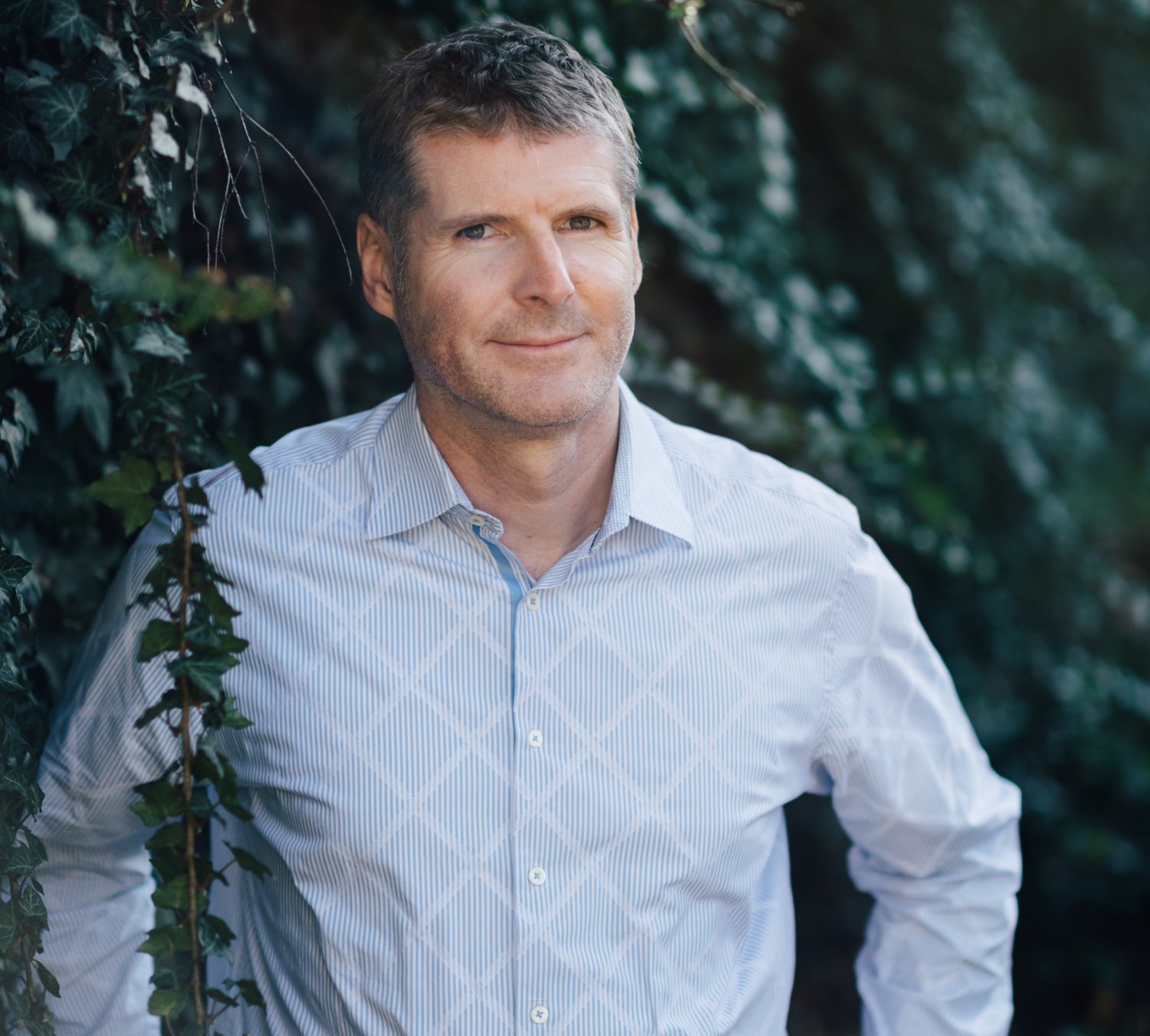 Portrait of Peder Olofsson in front of green bushes and dressed in a light-blue shirt.