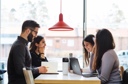 Fyra studenter sitter runt ett bord och samtalar. || Fyra studenter som sitter vid ett bord och studerar med sina laptops.