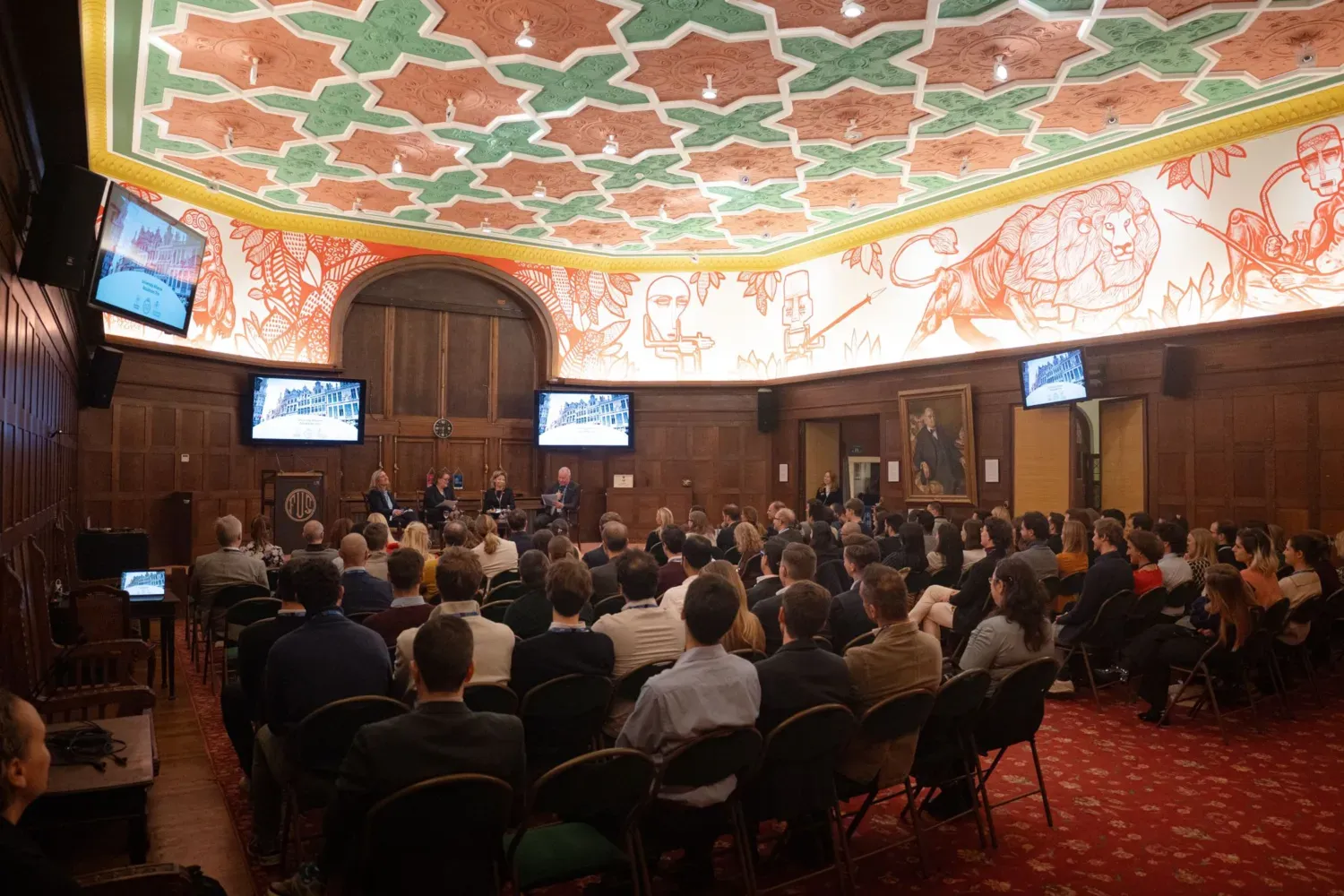 En publik sitter i en panmig sal, vänd mot en scen, där det sitter fyra personer med mickar.