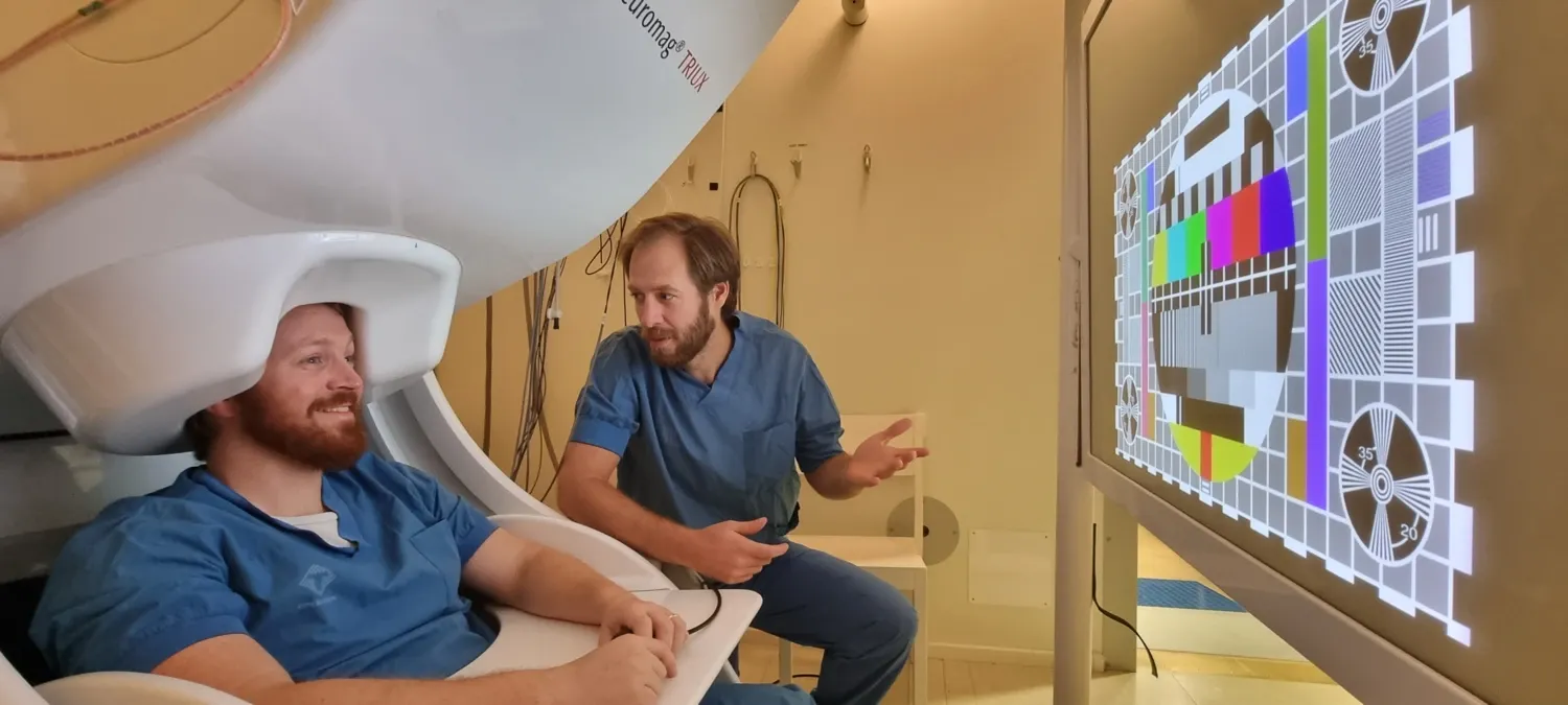 Two people are sitting in front of a large TV screen. One person is sitting under a MEG scanner