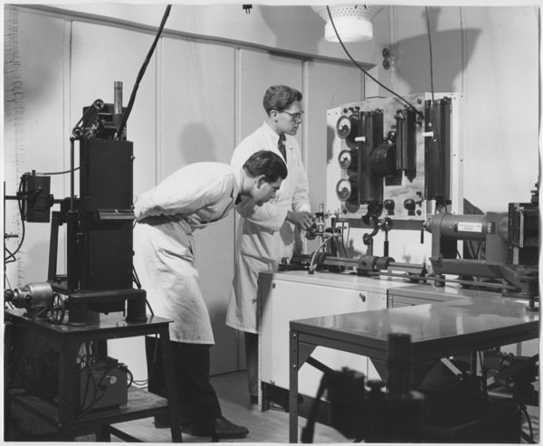 Men in lab coats by large measuring device.