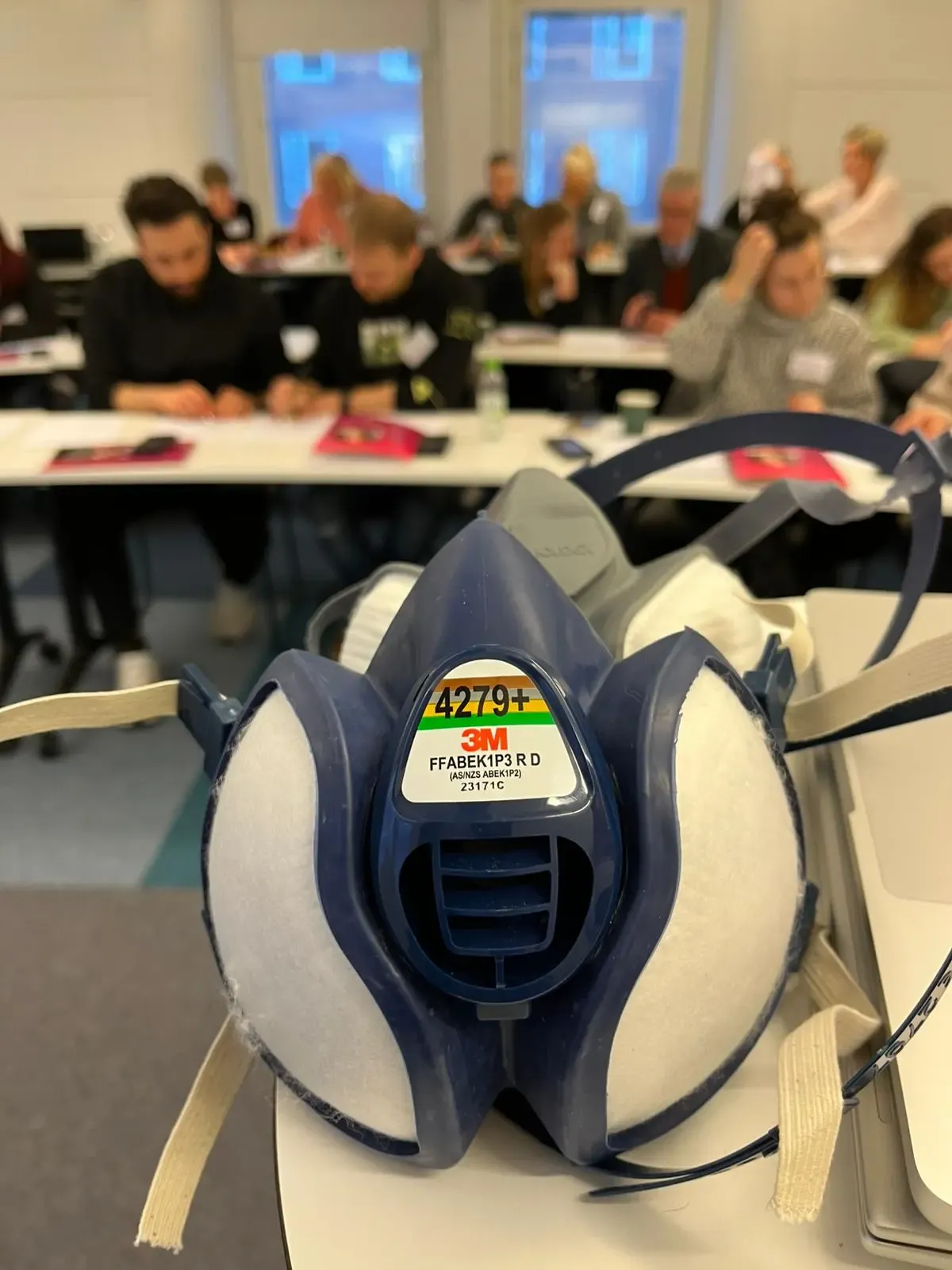 I förgrunden en skyddmask och i bakgrunden, i oskärpa, personer som sitter i en undervisningssal