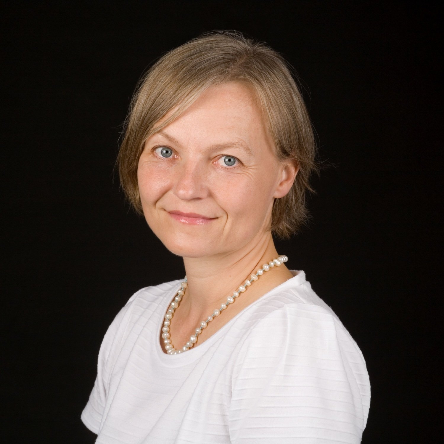 A woman wearing a white t-shirt and a white pearl neckless, standing in front of a black background.