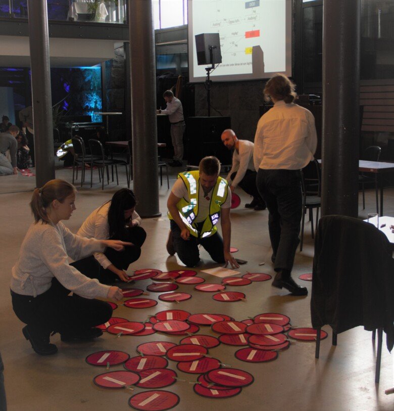 Four people stand or sit around a number of red tablets spread across the floor, in the background there are two more persons