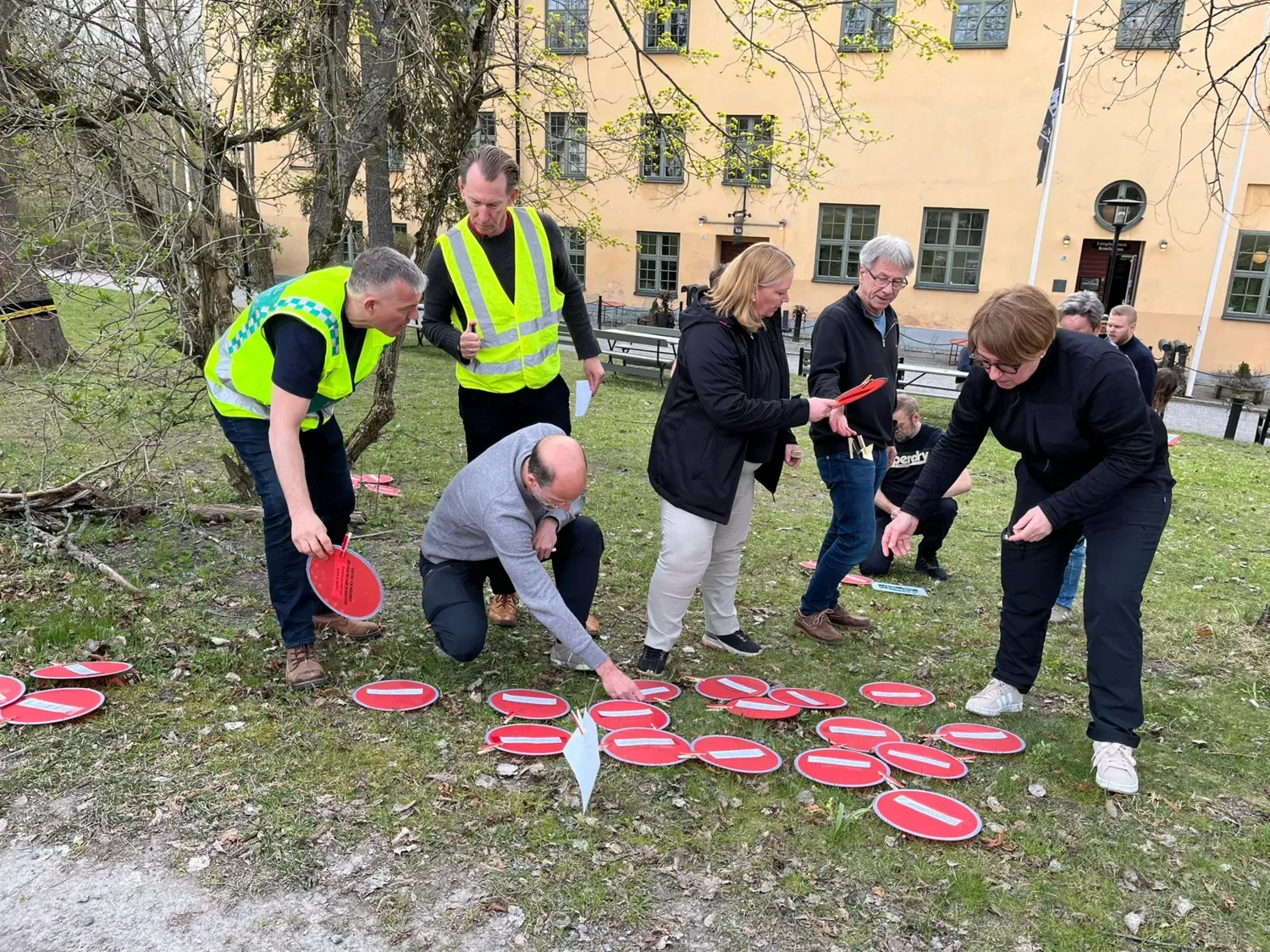 Flera personer, varav två bär reflexvästar, står utomhus på en gräsplan och sorterar röda frisbee-liknande plattor. I bakgrunden syns en gul byggnad