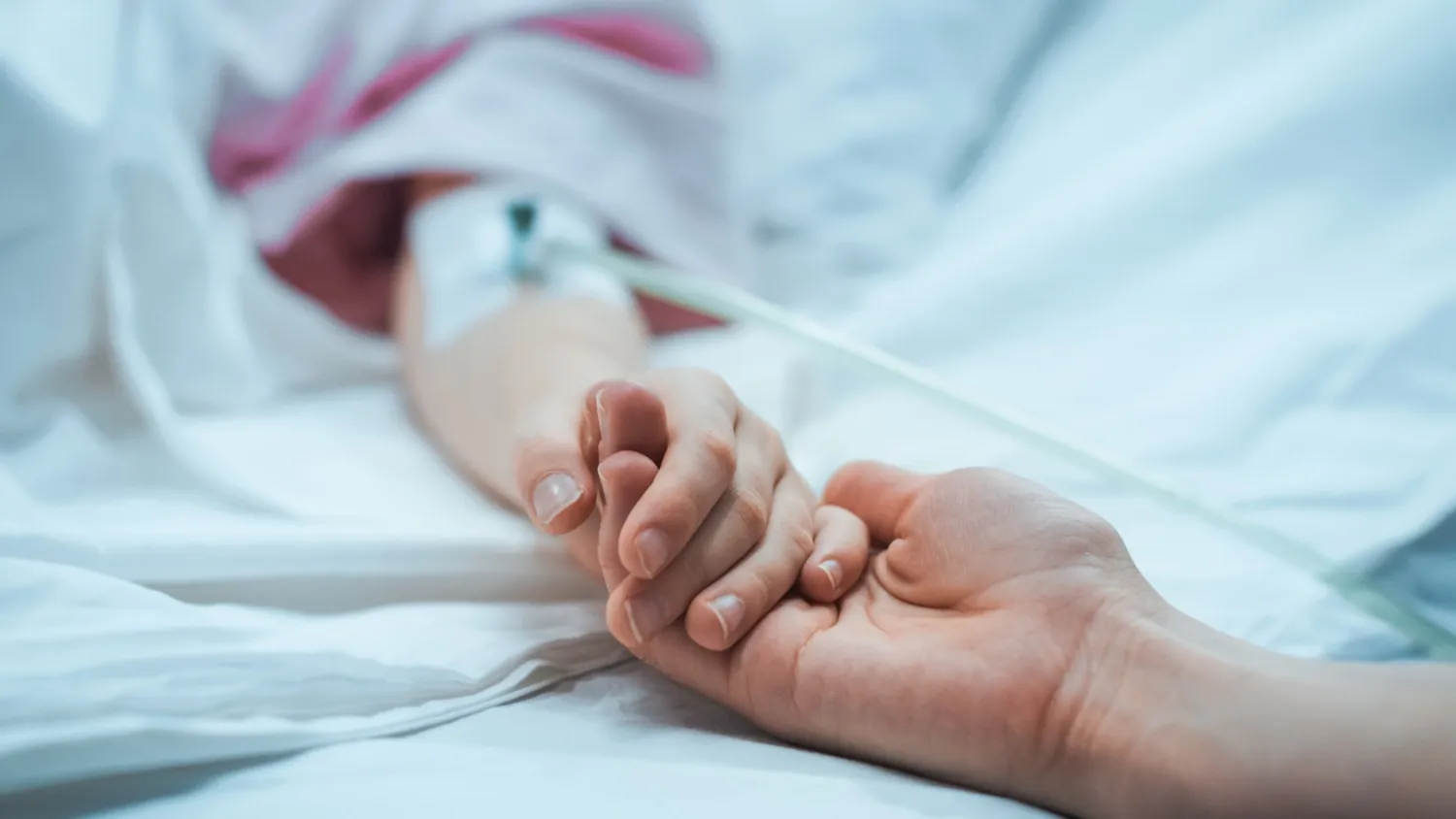 An adult's hand is holding a child's hand, and the child is lying in a hospital bed.