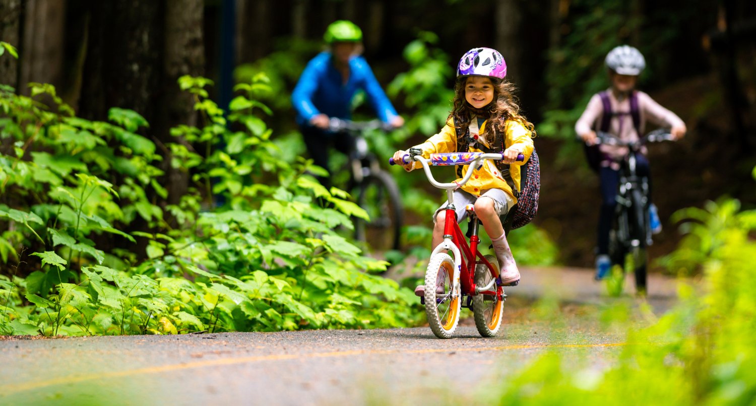 En liten flicka cyklar i skogen med sin mamma och ett syskon. Hon har cykelhjälp på sig.