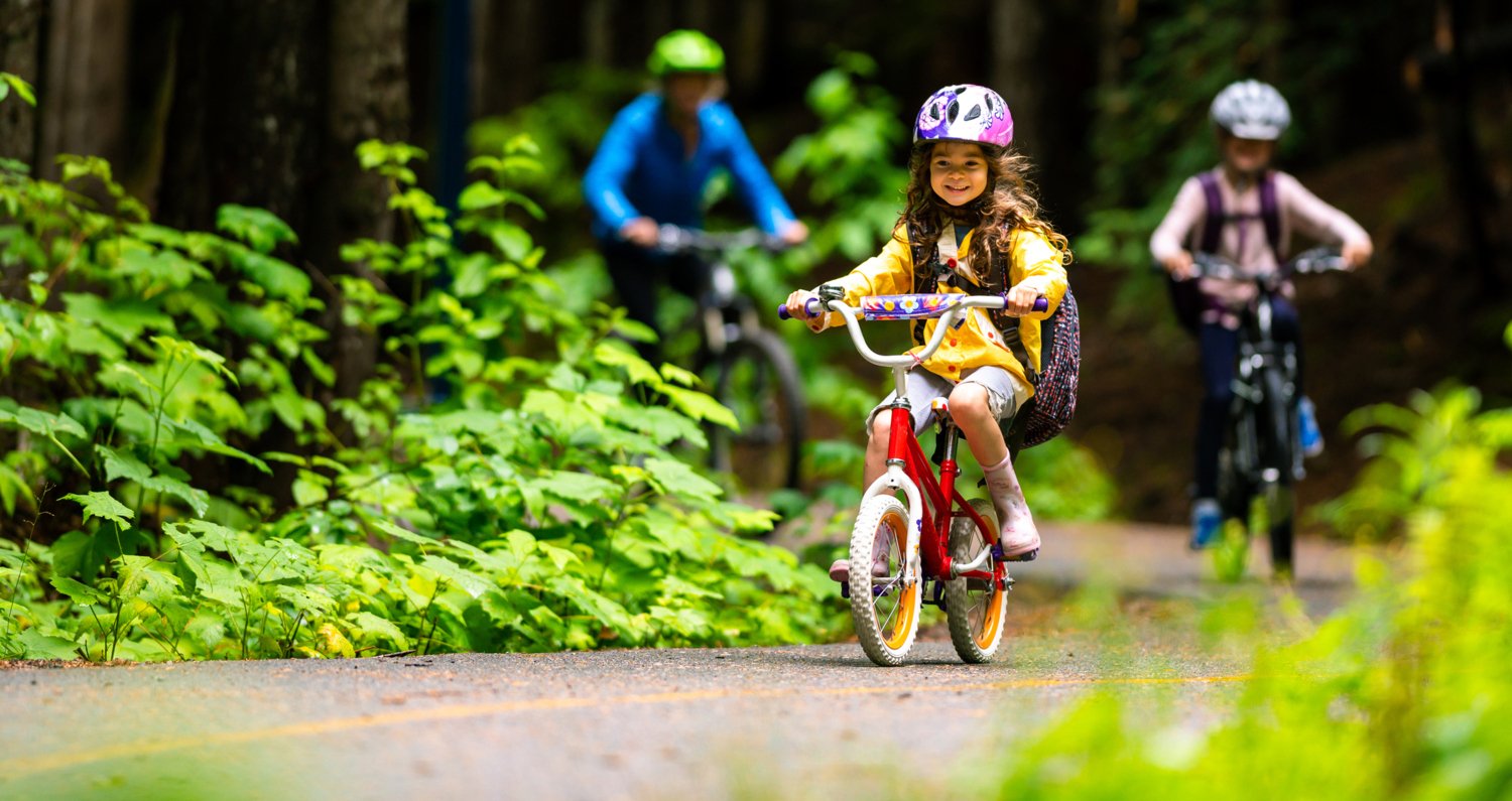 En liten flicka cyklar i skogen med sin mamma och ett syskon. Hon har cykelhjälp på sig.