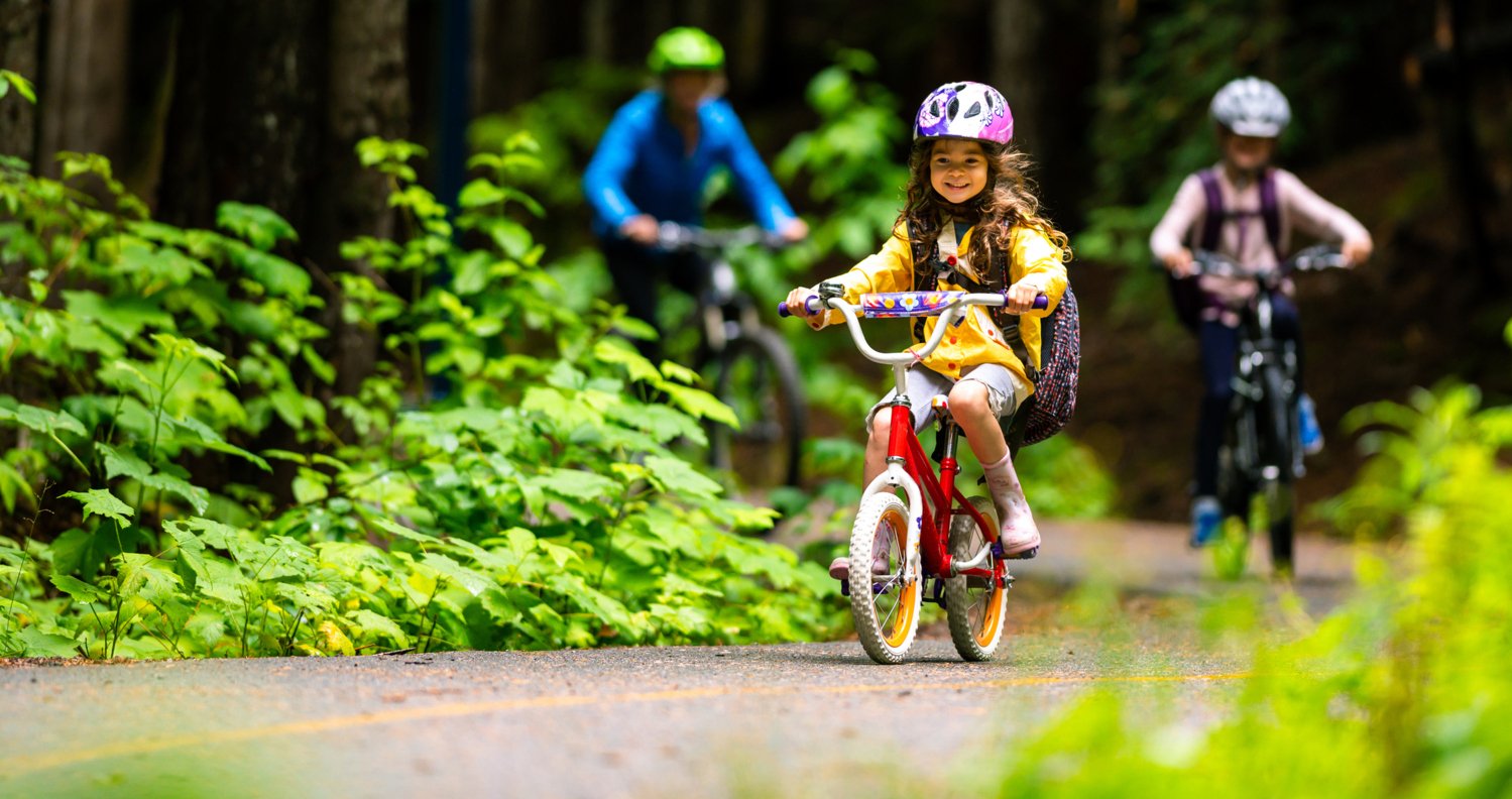 En liten flicka cyklar i skogen med sin mamma och ett syskon. Hon har cykelhjälp på sig.