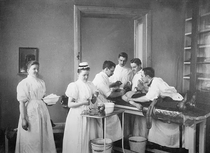 En patient ligger på ett operationsbord, några läkare står lutade över honom. I förgrunden arbetar två sjuksköterskor med förberedelser.
