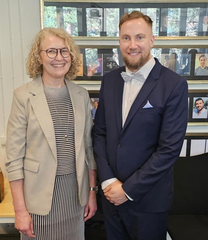 Opponent och doktorand framför en vägg av foton i ett konferensrum