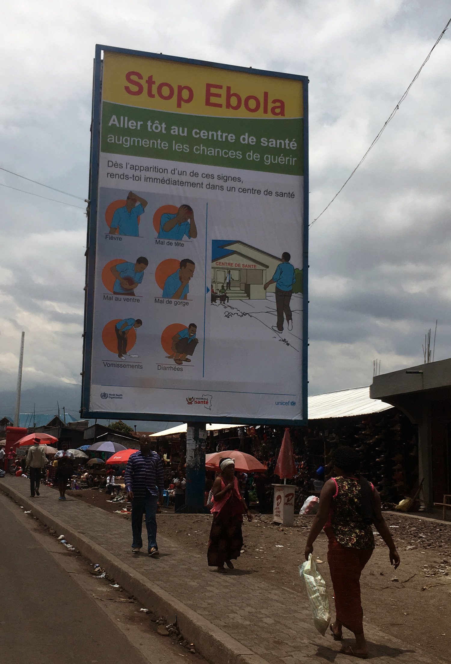 Sign with information about Ebola, along a street with cars, houses and people in the DR of Congo.