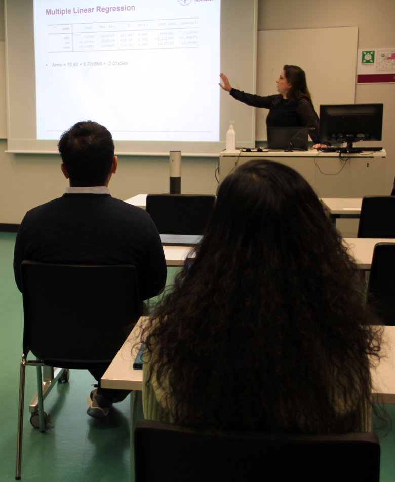 Två studenter, en man och en kvinna, sitter med ryggen mot kameran och tittar på en white board som deras lärare pekar på