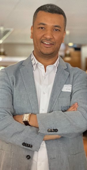 Portrait of a man, wearing a grey jacket, standing with his arms crossed.