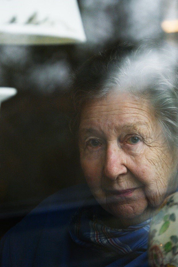 Porträtt av en äldre dam som tittar ut genom ett fönster.