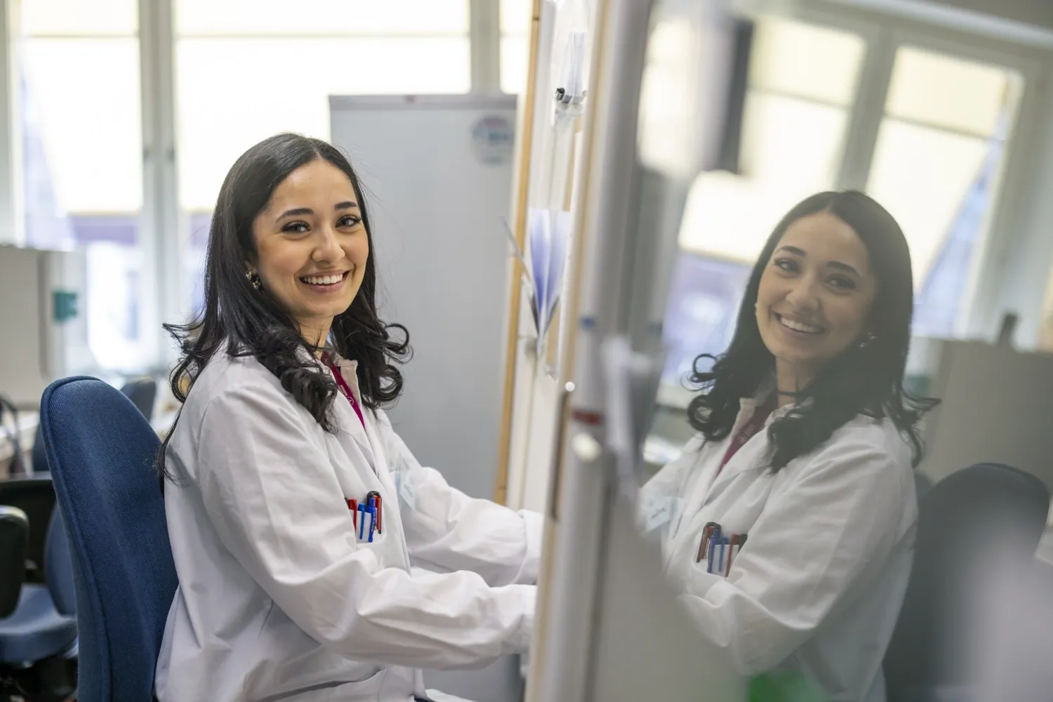 Kvinna i labbrock tittar in i kameran och ler. Hennes ansikte reflekteras i en glasdörr.