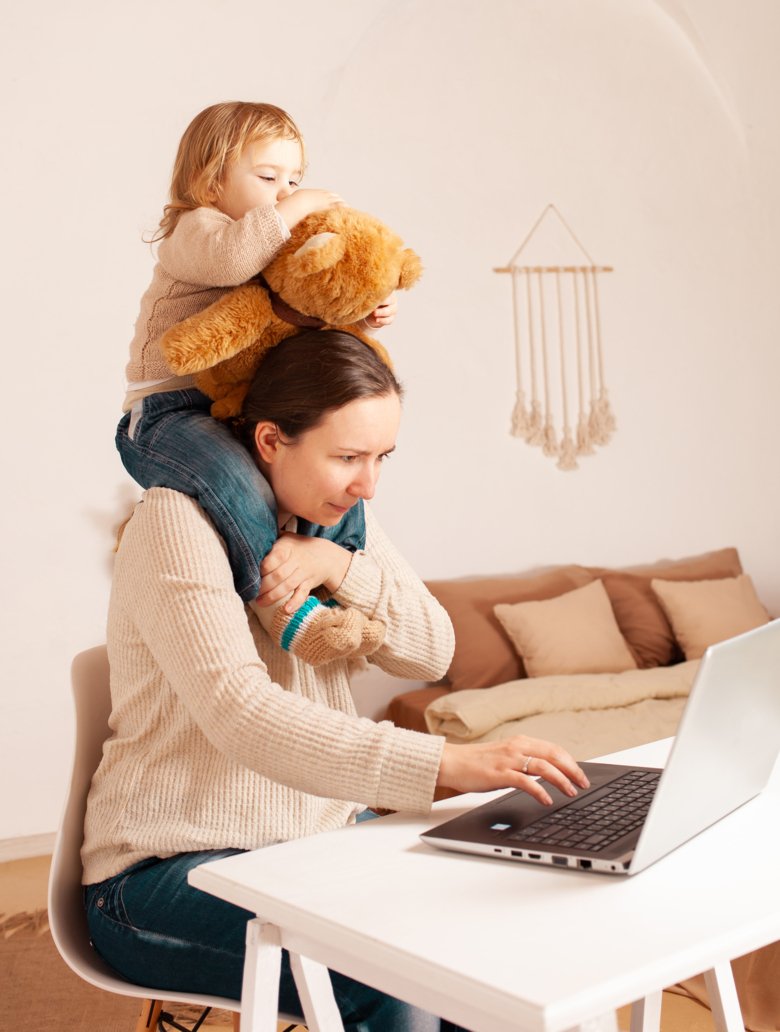 En mamma som sitter vid datorn och har dottern på axlarna.