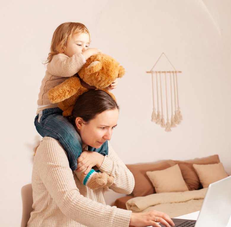 En mamma som sitter vid datorn och har dottern på axlarna.