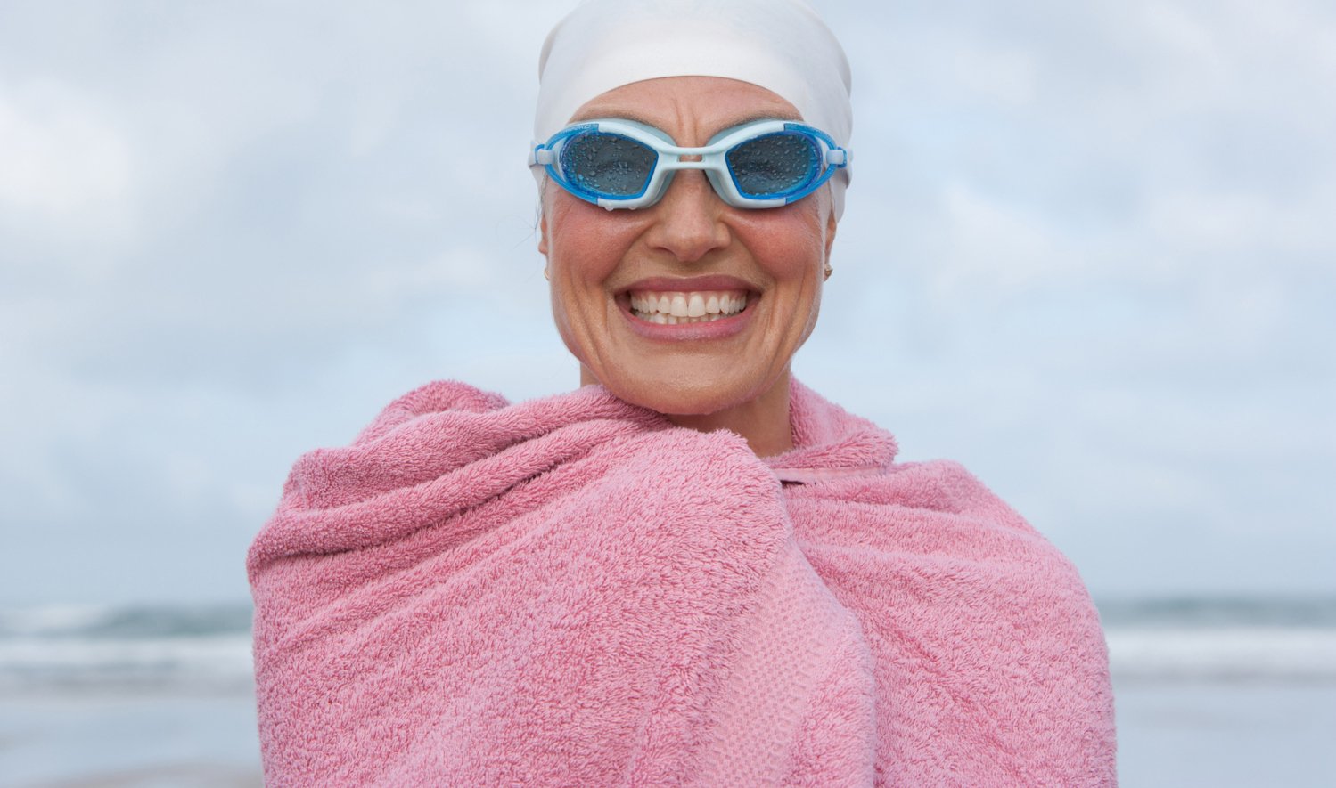 Glad kvinna med simglasögon och badlakan över axlarna har just kommit upp ur havet.