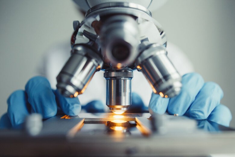 Microscope with light and researcher wearing blue gloves standing behind it.