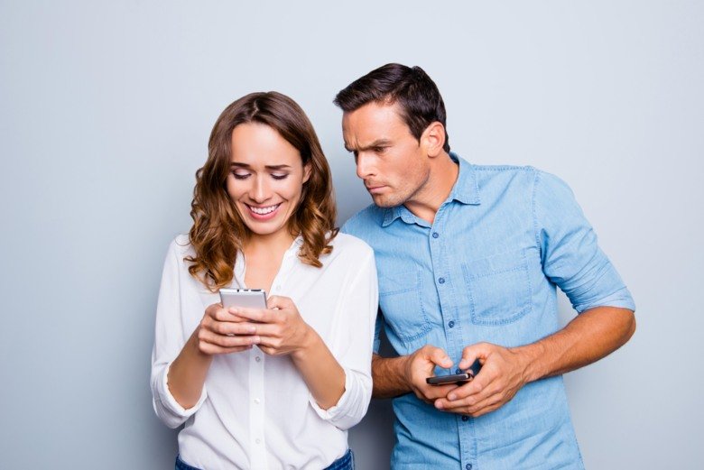 Picture of a man looking at the woman's phone over her shoulder and getting jealous.