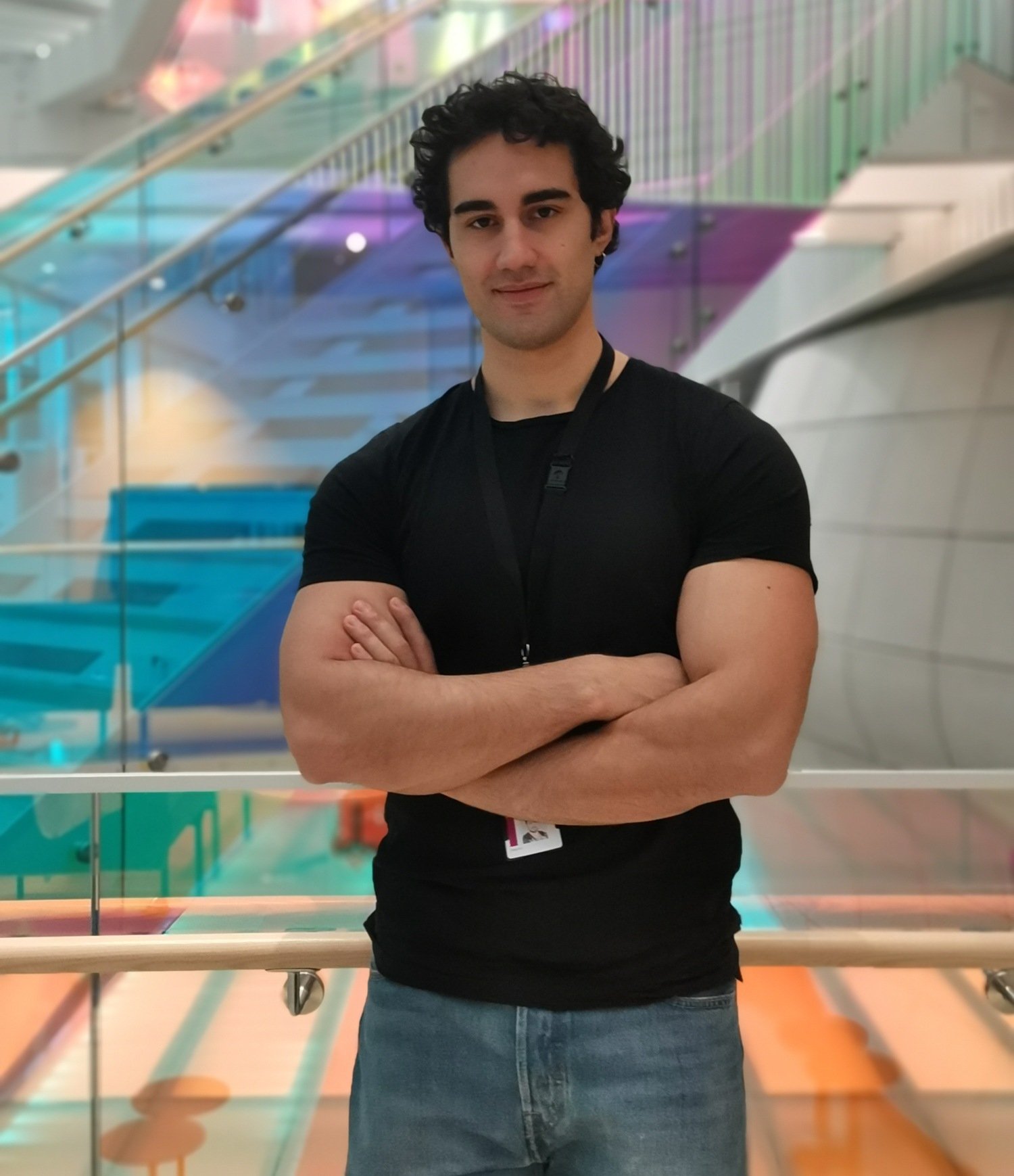 A man wearing a black T-shirt and jeans. He is standing with his arms crossed and is photographed indoors.