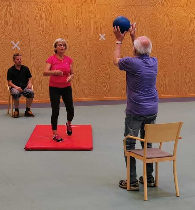 Personer i ring, en del på stolar, andra som står upp. De kastar en boll till varandra.