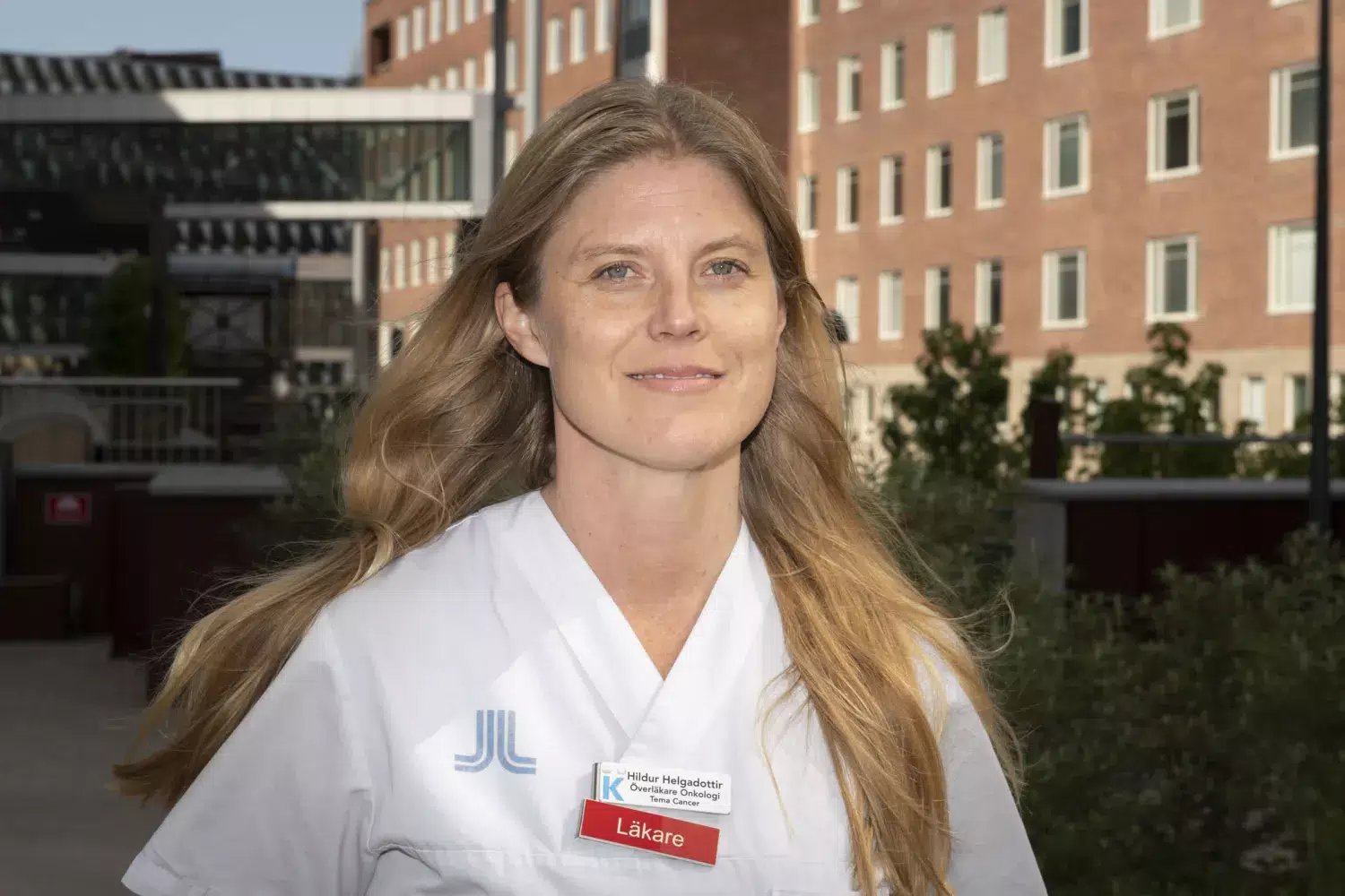 Porträtt av Hildur Helgadottir iklädd sjukhuskläder. Hon har långt ljust hår och står utomhus.