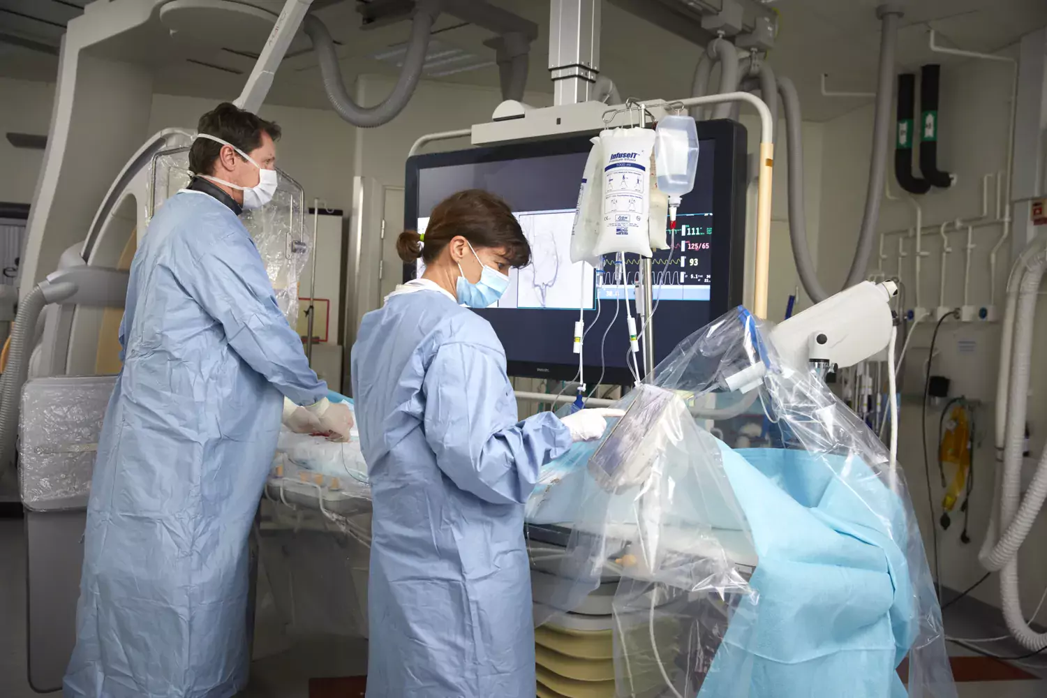 Två personer i operationskläder med dropp, skärm och annan medicinsk utrustning. De arbetar och har ryggen mot kameran.