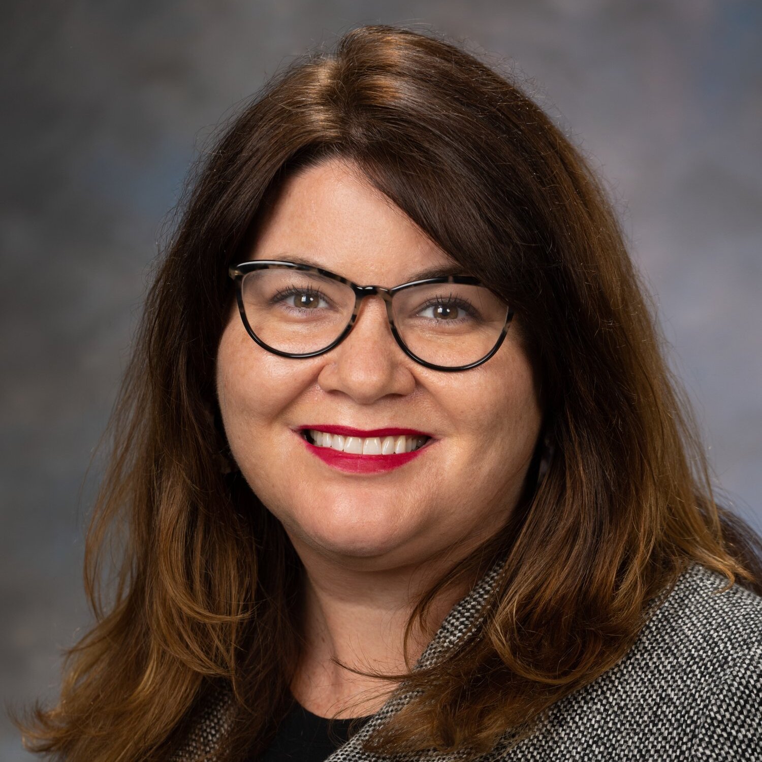 A woman with dark brown hair and glasses, wearing a knitted sweather, sitting in front of a dark grey background.