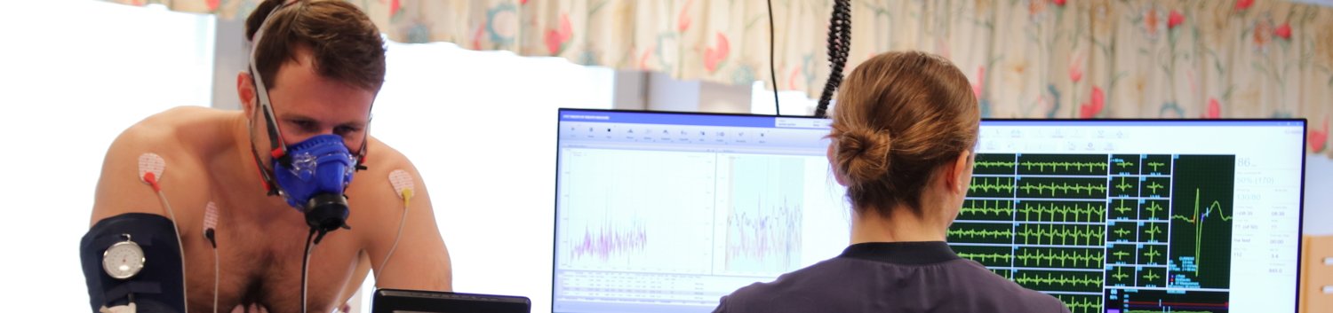 A person does a work test on a test bike. The person has a breathing mask for gas analysis and ECG electrodes attached to them. A test supervisor monitors the test and looks at a screen showing different curves of the test.