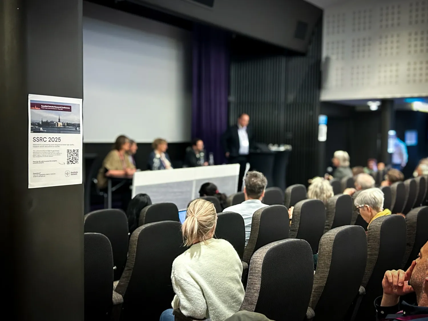 Åhörare i en föreläsningssal. På scen sitter en panel. Fokus ligger på en affisch på en stolpe i rummet.