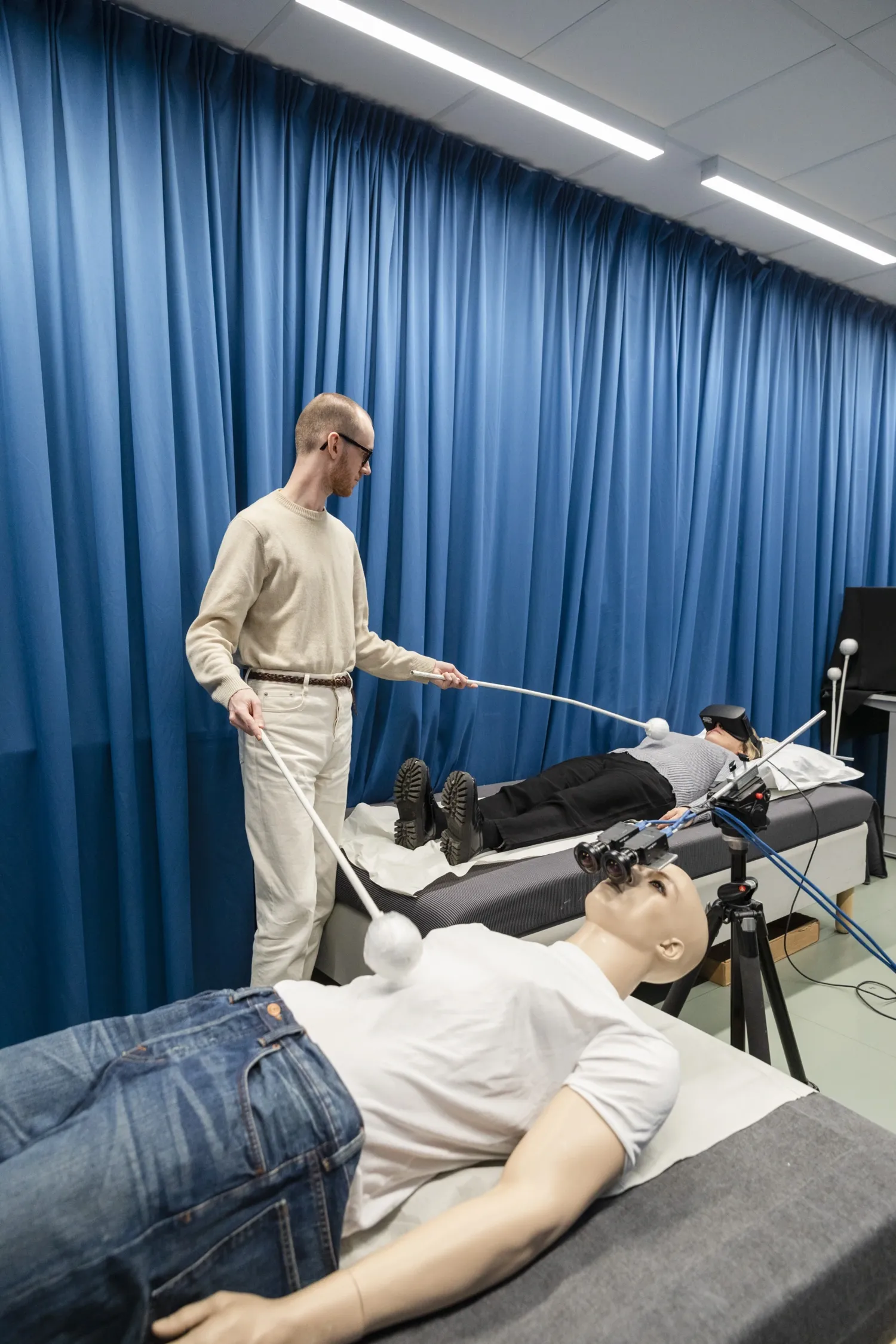 A mannequin lies on a cot in the foreground and a person lies on a cot in the background. A person stands up and touches both the mannequin and the person during the experiment.