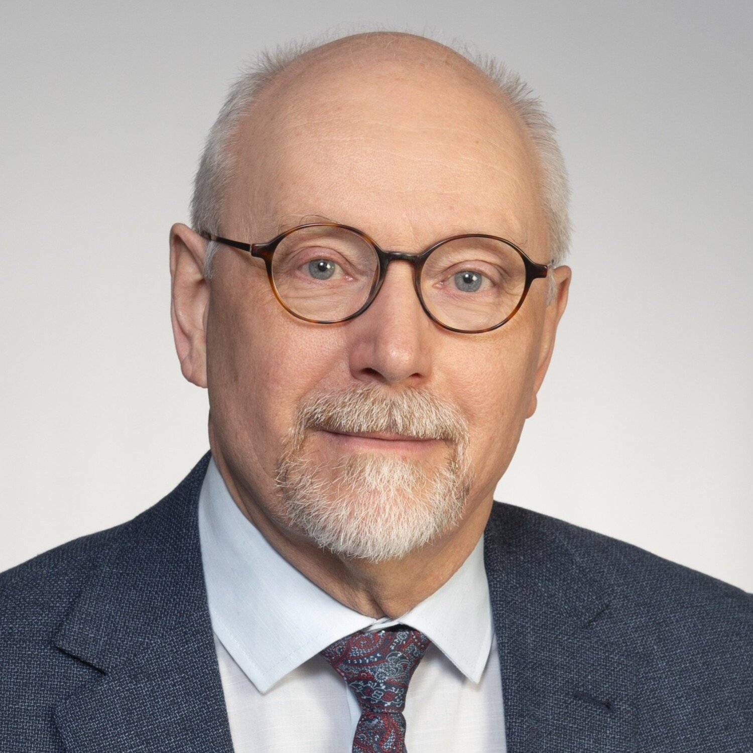 A portrait of a man who wears a suit, tie and glasses, sitting in front of a gray background.