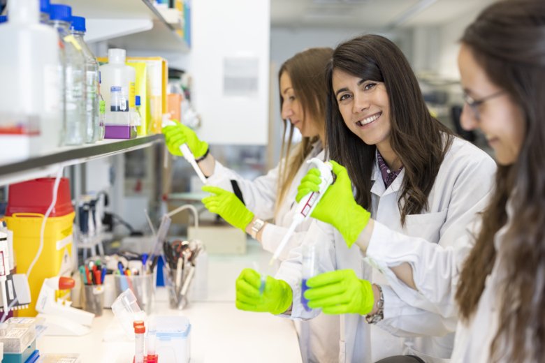 Tre kvinnliga forskare står med provrör och pipetter i labbmiljö. En av dem tittar in i kameran.