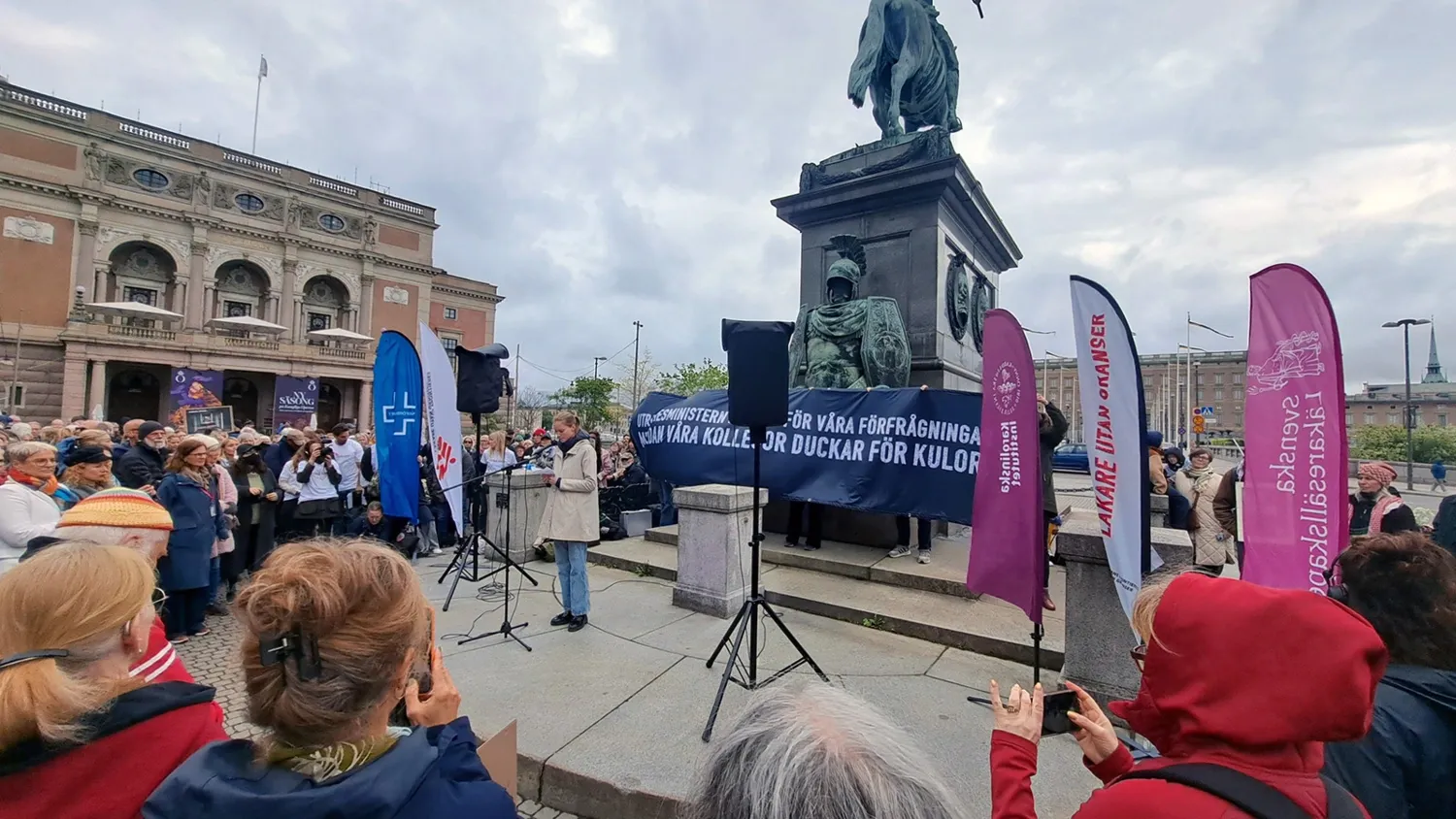 Ett antal människor är samlade på torget. Där finns en scen där Caroline läser upp vittnesmål. På scenen finns banderoller som visar vilka som är arrangörer.