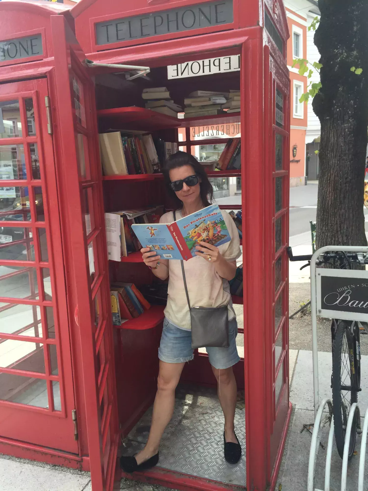 Marie Carlén in a telephone booth, reading a magazine. The telephone booth is a library filled with books.