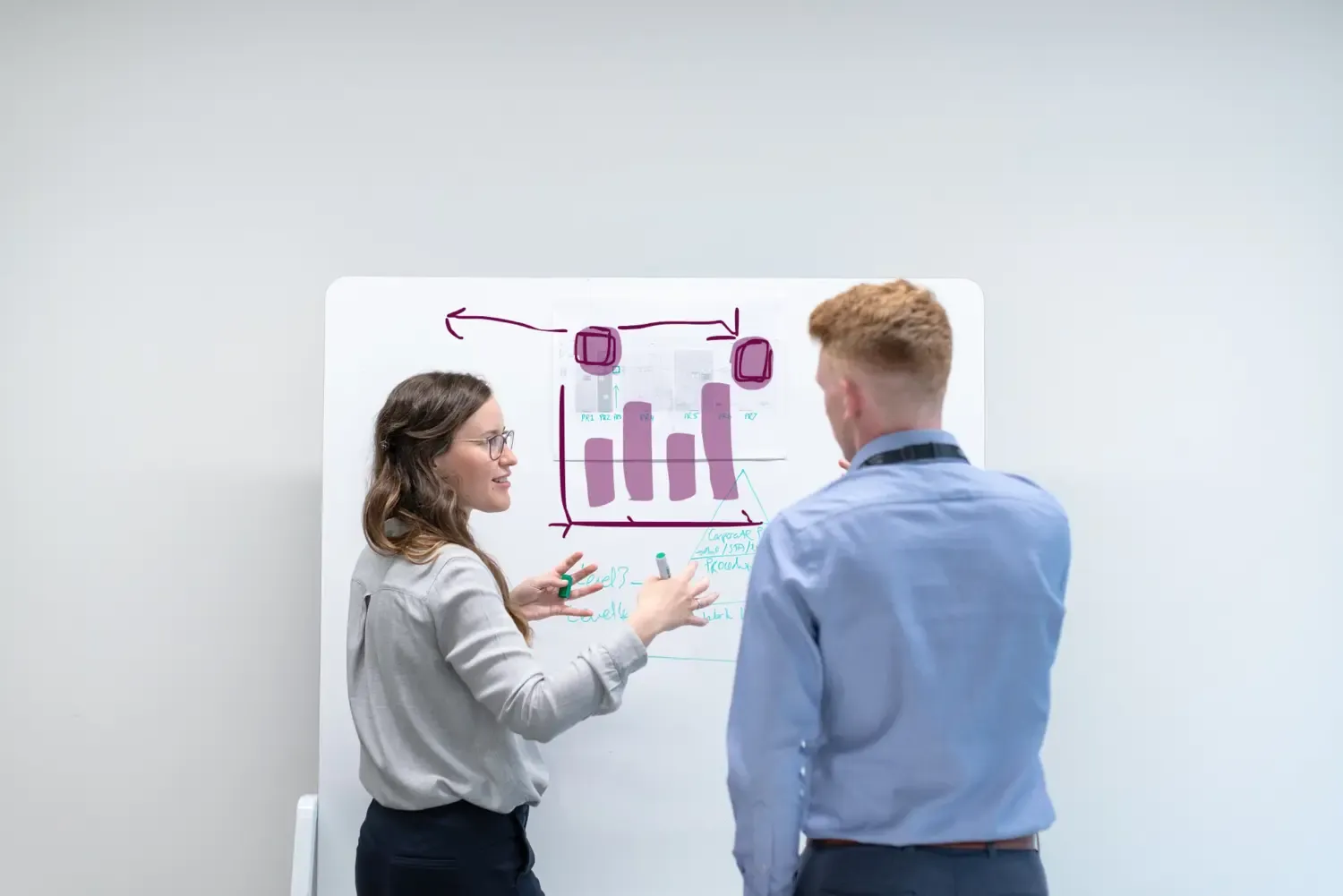 Two people standing in front of a white board discussing and writing.