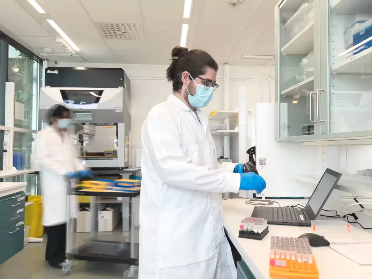 Två personer i vita labbrockar står i ett labboratorium. Den ene scannar ett provrör
