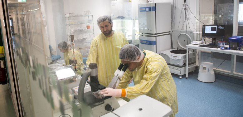 One scientist is looking in a microscope another scientist is standing next to him. Both have yellow clothes.