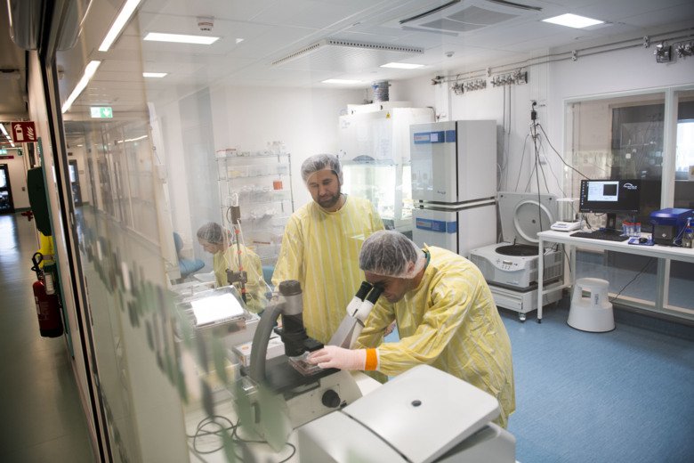 One scientist is looking in a microscope another scientist is standing next to him. Both have yellow clothes.