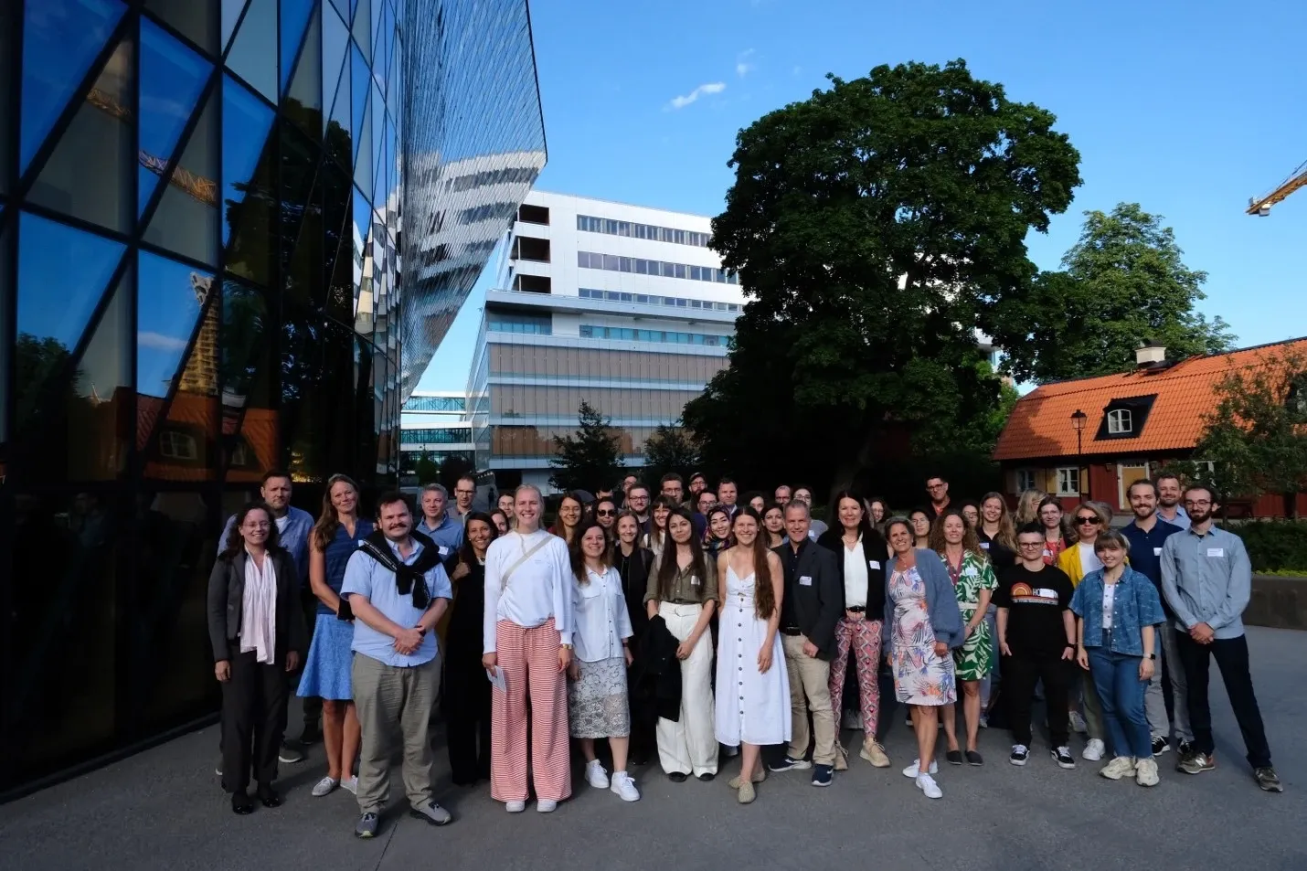 Group picture of about fifty people gathered outdoors at KI campus Solna. To the left is a modern glass facade. To the right is an older red wooden building.