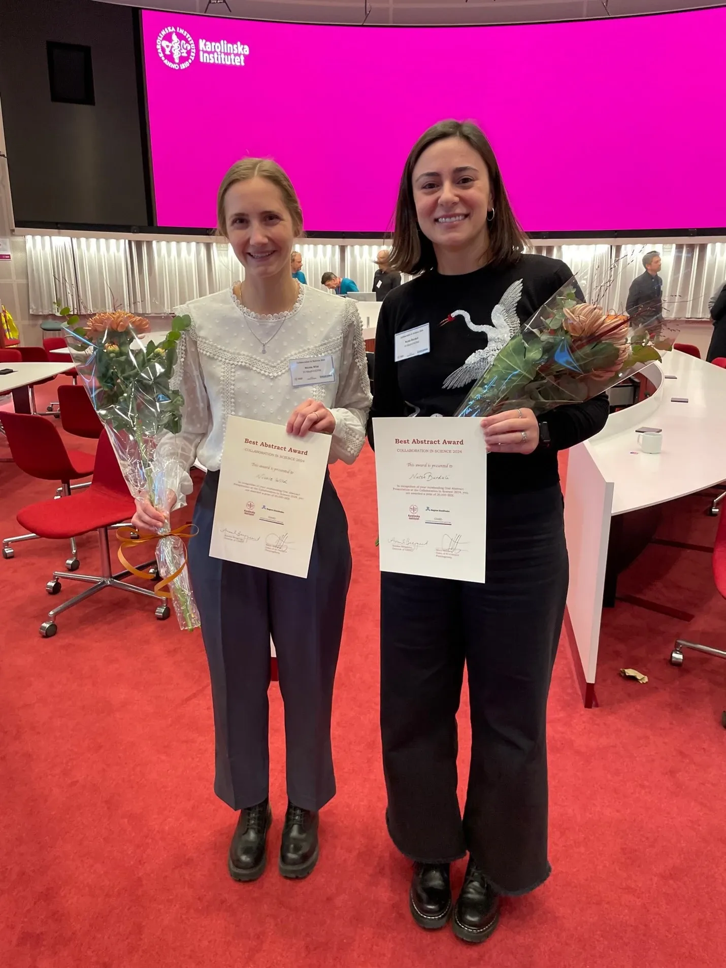 Two women stand side by side, each holding a diploma and a bouquet of flowers.