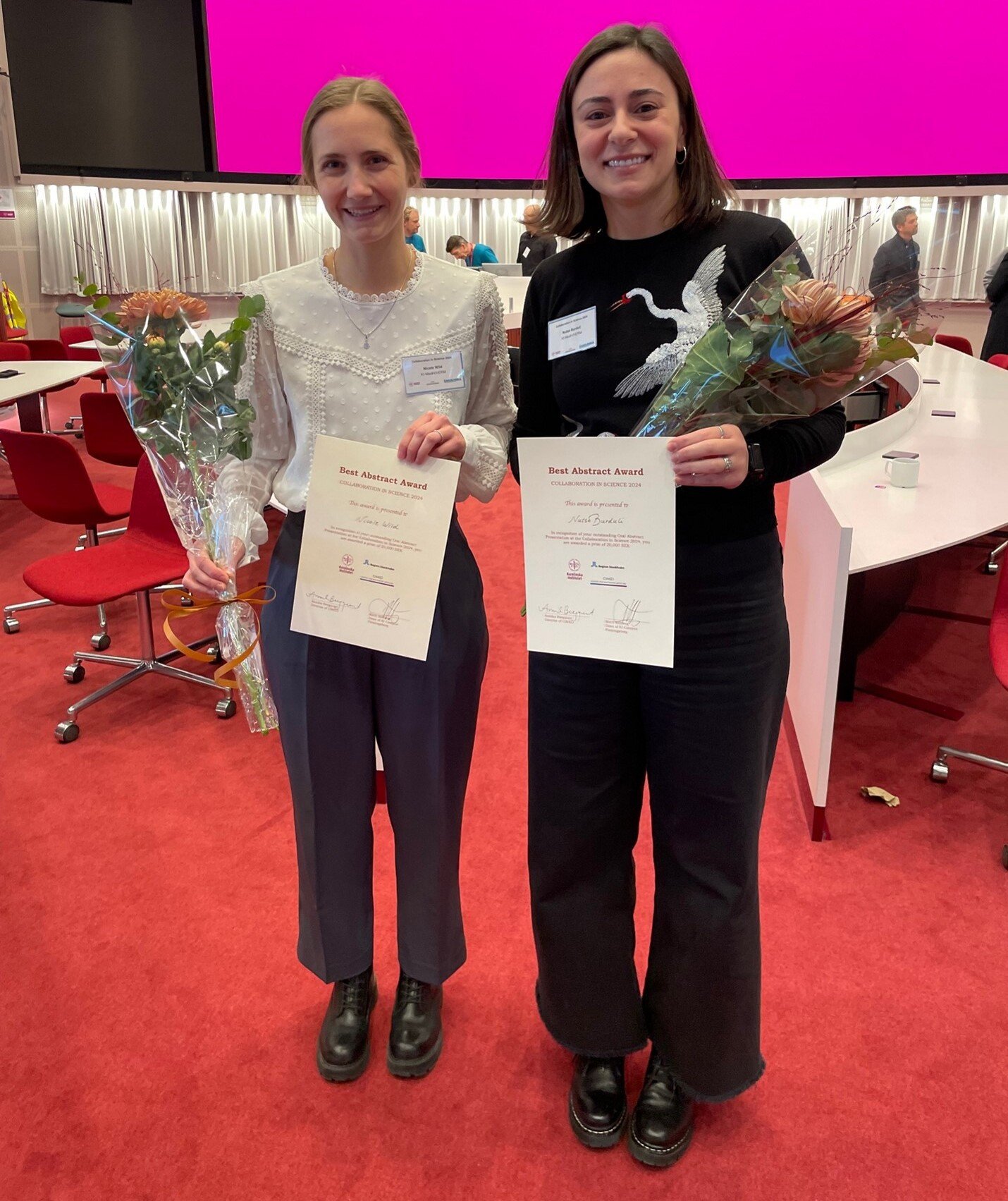 Two women stand side by side, each holding a diploma and a bouquet of flowers.