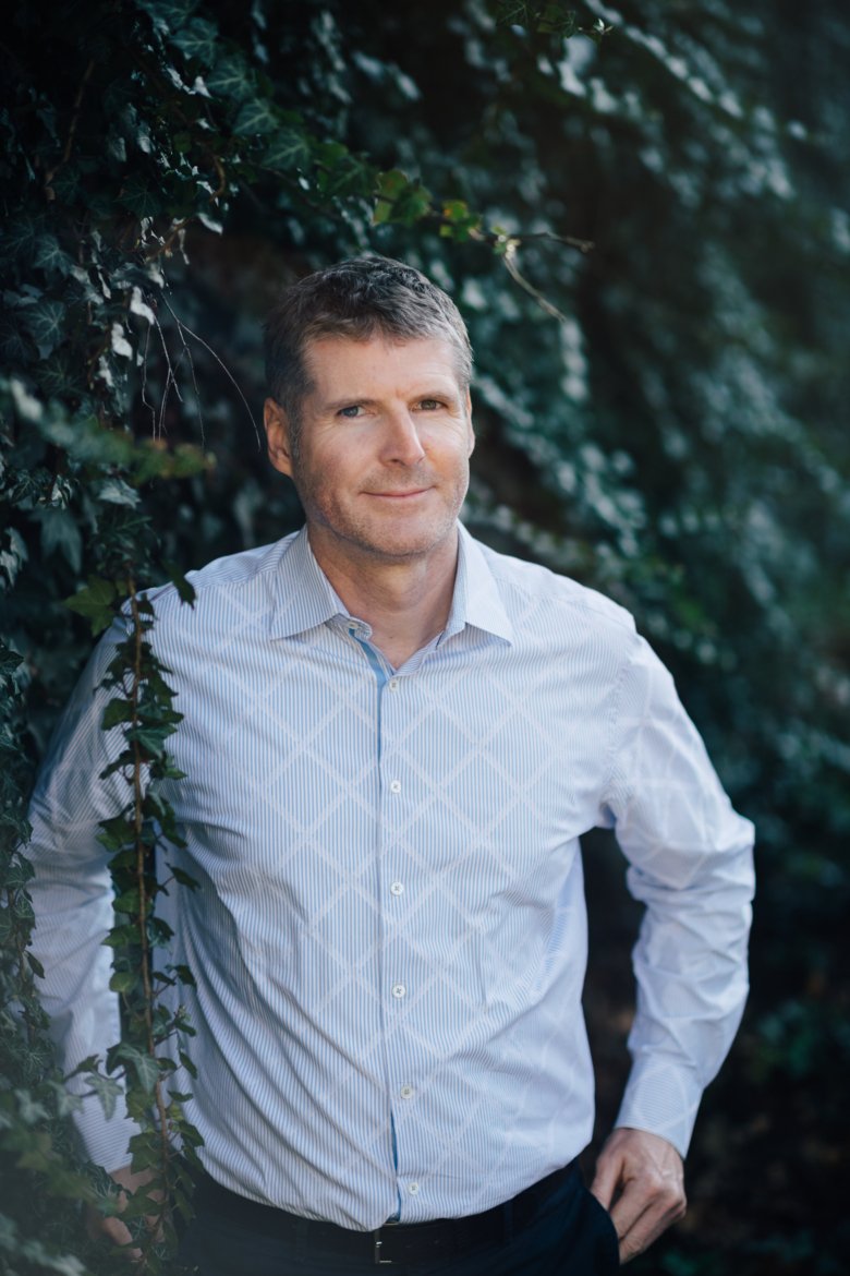 Porträtt av Peder Olofsson framför ett buskage och iklädd ljusblå skjorta.