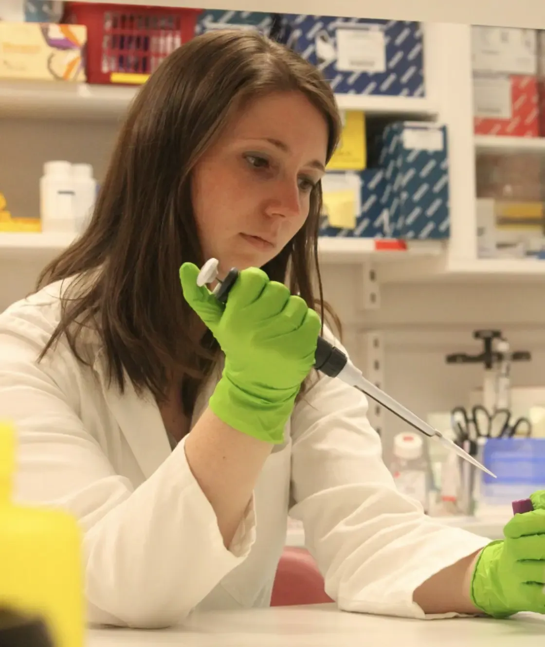 A woman in a white lab coat and green rubber gloves handles a sample.