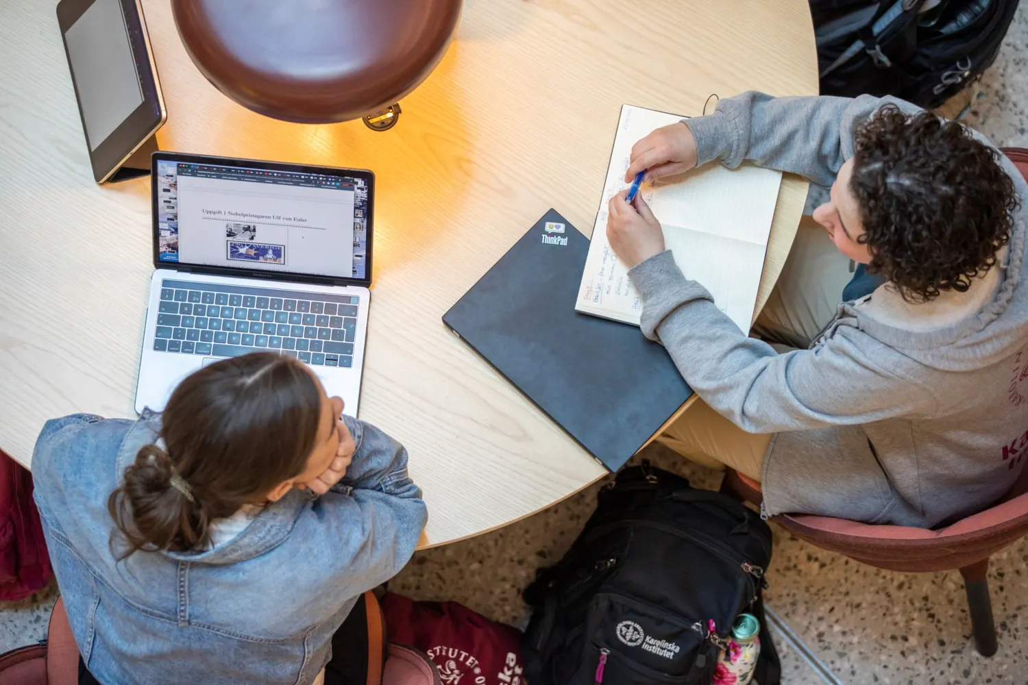 Studenter inomhus på campus Solna vårterminen 2022. || Studenter som umgås inomhus på campus. || Två studenter sitter vid ett bord och samtalar