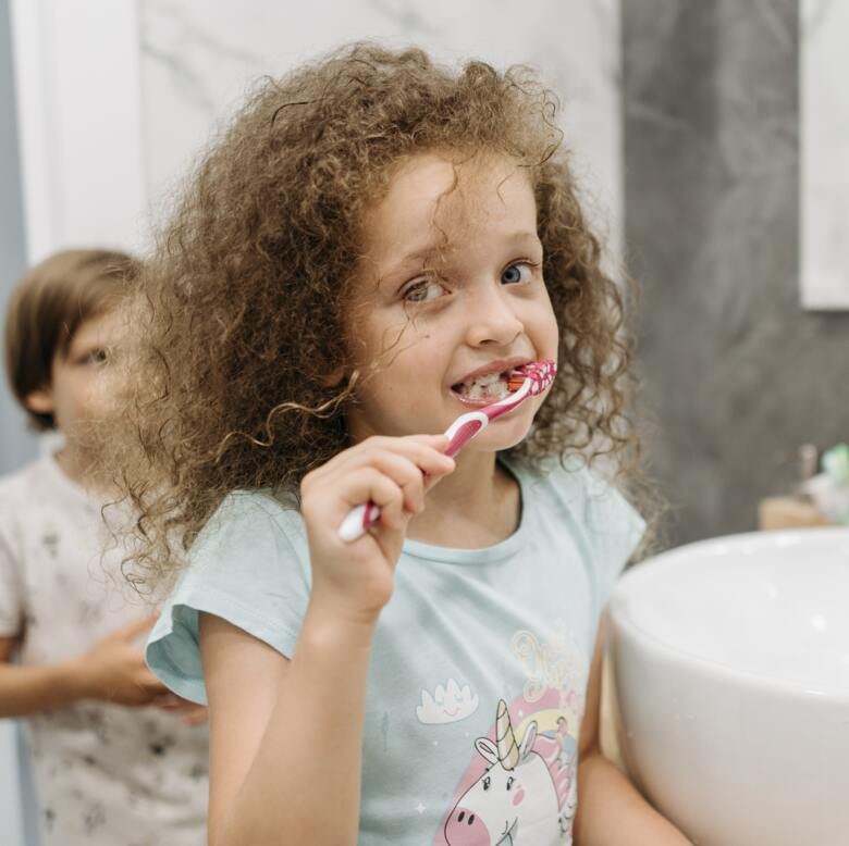 En liten flicka med brunt lockigt hår står framför spegel i badrum och borstar sina tänder